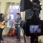 A man and a woman playing guitars in front of a camera