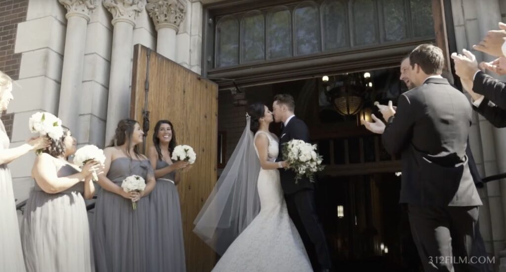The bride and groom are kissing in front of St. Michael's Church in Chicago, and all the guests are cheering.