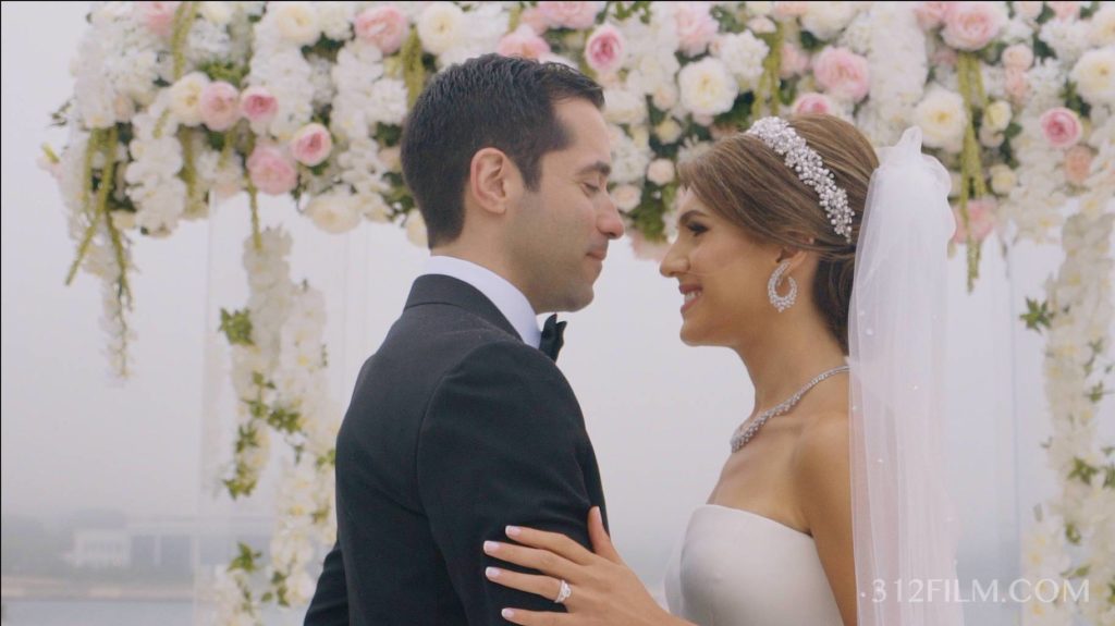 The bride and groom are standing by a flower arch at the Adler Planetarium in Chicago, ready for their ceremony. The video was filmed by 312 Films and the photos were taken by Clary Pfeiffer Photography.