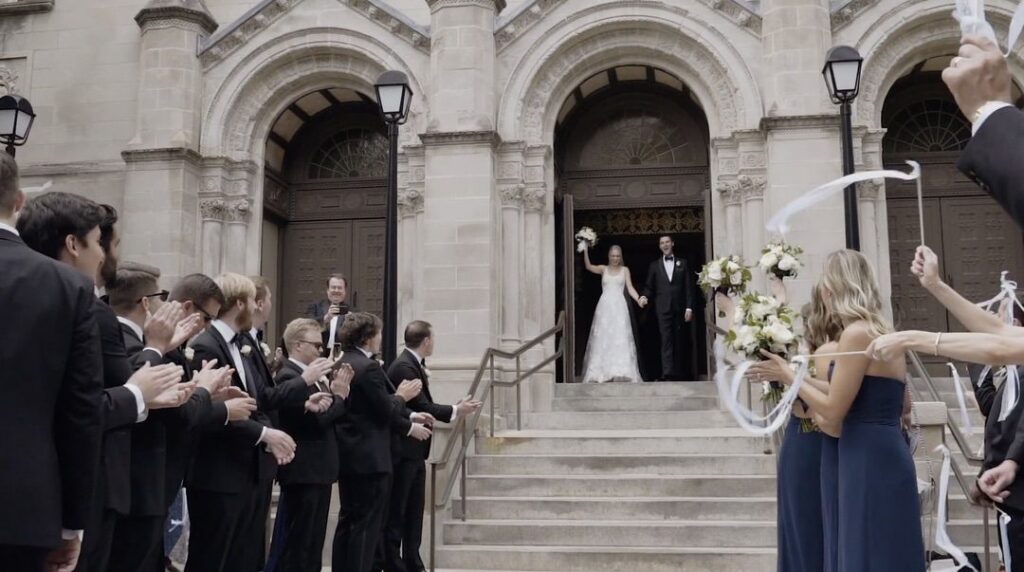 The bride and groom are leaving Saint Clement Church in Chicago after the ceremony, with their guests cheering them on outside.