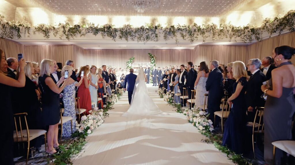 The bride is walking down the aisle with her father, with the flower decorations from Steve's Flower Market in the background.